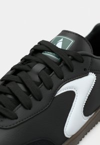 Basket noire avec un détail blanc sur le côté et des lacets noirs, avec une étiquette verte comportant un logo blanc sur la languette.