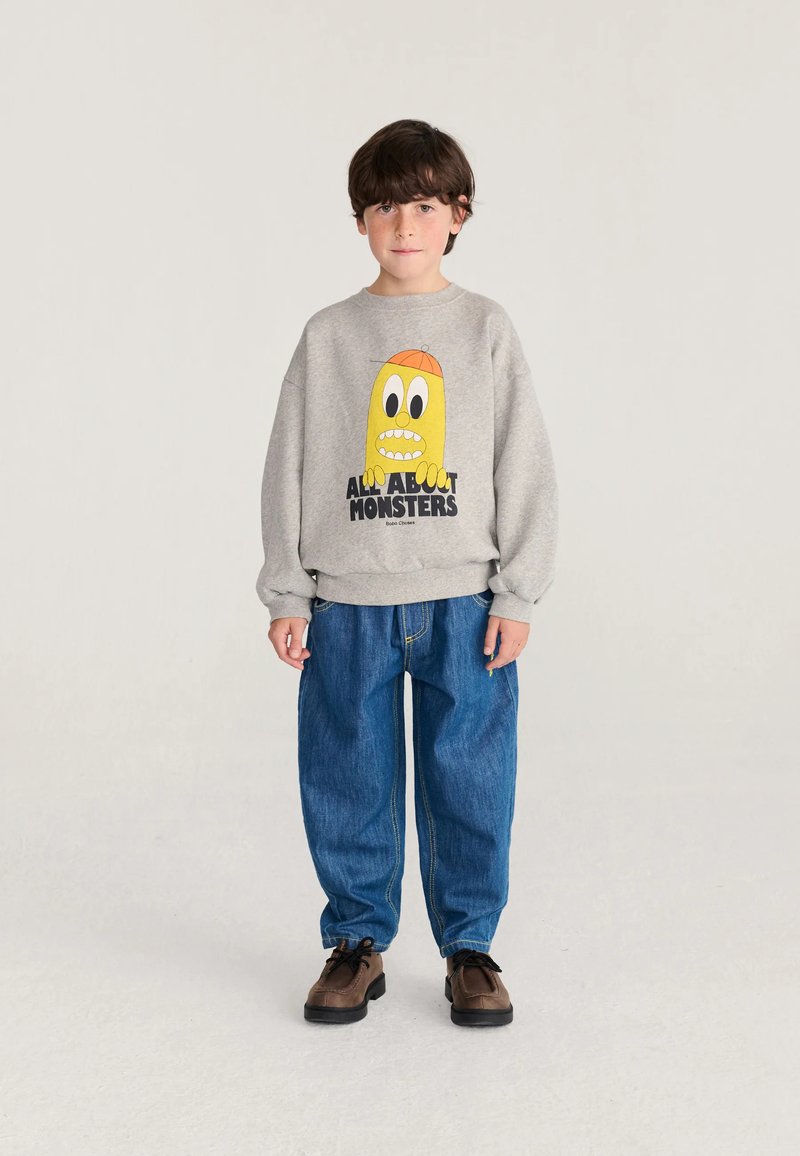 Ragazzo in piedi che indossa una felpa grigia con un mostro giallo dei cartoni animati e jeans blu su sfondo chiaro uniforme.