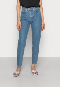 Jeans de ganga azul de cintura alta com pernas ajustadas. Apresenta cinco bolsos e fecho de botão. Combinado com sandálias brancas.