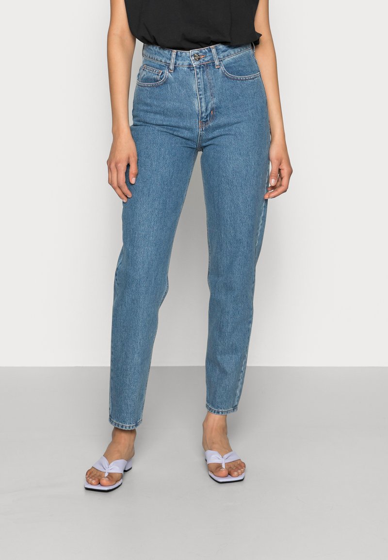 Jeans de ganga azul de cintura alta com pernas ajustadas. Apresenta cinco bolsos e fecho de botão. Combinado com sandálias brancas.