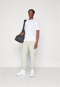 Chemise de sport blanche à manches courtes, pantalon cargo beige clair et baskets blanches, avec un sac de sport noir tenu à la main. Design minimal, look sportif.