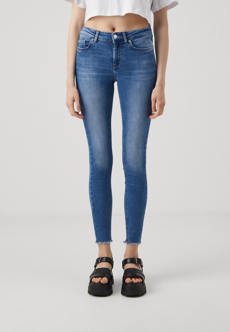 Jeans de denim azul ajustados con cintura alta, dobladillo deshilachado en el tobillo y bolsillos frontales, combinados con sandalias de plataforma negras.