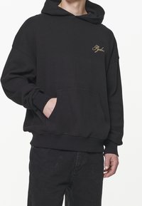 Sudadera con capucha negra en tejido texturizado, que cuenta con un bolsillo frontal y un logo bordado en dorado. Corte holgado con puños y bajo acanalados.