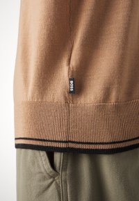 Beiger Strickpullover mit geripptem Saum, ausgestattet mit einem schwarzen Streifen und einem seitlichen Etikett mit der Aufschrift "BOSS". Kombiniert mit olivgrünen Hosen.
