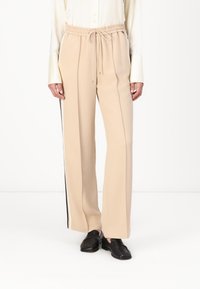 Beige wijde broek met een elastische tailleband, trekkoorddetail en zwarte zijstrepen. Glad, zacht materiaal met een comfortabele pasvorm.