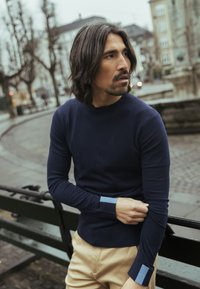 Tumši zils piegulošs džemperis ar garām piedurknēm, ar gaiši zilām detaļām manšetēs. Mīļa tekstūra, apaļa kakla izgriezuma, valkāts ar bēšām biksēm.