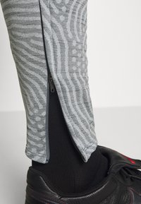 Primer plano de una pierna inferior que lleva pantalones grises con un patrón y con la cremallera lateral parcialmente abierta, un calcetín negro y una zapatilla negra con costuras rojas.