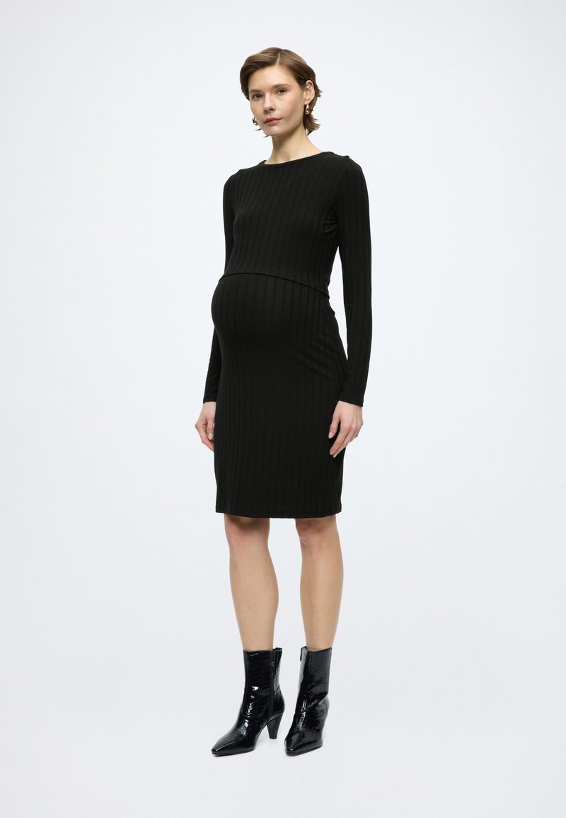 Μαύρο ριμπ φόρεμα εγκυμοσύνης με μακριά μανίκια και εφαρμοστή σιλουέτα, σε συνδυασμό με λαμπερά μαύρα μποτάκια μέχρι τον αστράγαλο. Απλός και κομψός σχεδιασμός.