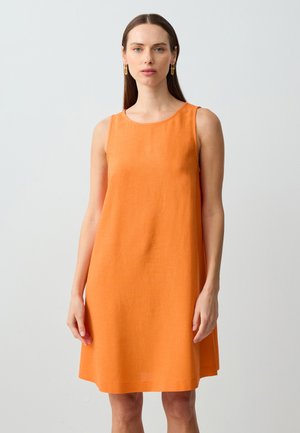 Donna con capelli castani lisci, che indossa un abito arancione brillante senza maniche, lungo fino al ginocchio, e orecchini a cerchio dorati, in piedi su uno sfondo chiaro e semplice.