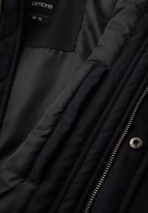 Interior de chaqueta negra, tejido suave con textura acolchada, detalles visibles de cremallera y botones a presión, etiqueta de tamaño colocada en la esquina superior izquierda.