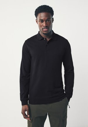 Tânăr cu păr scurt și creț, purtând un tricou polo negru cu mâneci lungi și pantaloni cargo verde măsliniu, stând pe un fundal simplu alb.