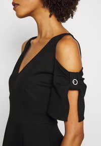Femme portant une robe noire à épaules dénudées avec une boucle circulaire décorative sur la manche et un décolleté en V.