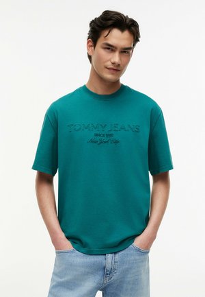 Junger Mann trägt ein türkisfarbenes "Tommy Jeans" T-Shirt und hellblaue Jeans, steht mit den Händen in den Taschen vor einem einfarbigen weißen Hintergrund.