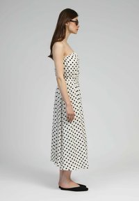 Robe sans bretelles, longueur cheville, de couleur blanche avec des pois noirs. Taille élastiquée, tissu lisse, portée avec des chaussures plates noires.
