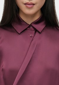 Bordowa jedwabna bluzka z klasycznym kołnierzykiem, z zapięciem na guziki i gładką fakturą. Materiał miękko układa się wokół dekoltu.