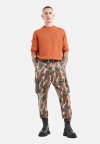 Oranger gestrickter Pullover mit Rundhalsausschnitt, kombiniert mit grünen, braunen und orangefarbenen Camo- Cargo-Hosen mit seitlichen Taschen und elastischen Bündchen. Schwarze Kampfstiefel.