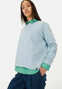 Jasnoniebieska bluza z ribbami na mankietach, noszona na zielonej koszuli z kołnierzykiem. Bluza ma subtelne logo i luźny krój.