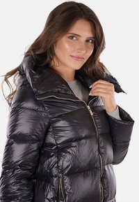 Giacca a vento nera con finitura lucida, colletto alto e dettagli della cerniera dorati. Presenta un design trapuntato e tasche con zip sul davanti.