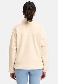 Cream fleece pullover with a high collar and dropped shoulders, showing a smooth texture. Paired with light blue pants, back view.