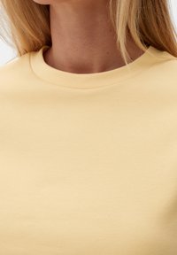 Detailní záběr na osobu, která má na sobě světle žluté tričko s kulatým výstřihem, viditelný krk, čelist a blond vlasy.