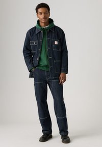 Jovem vestido com uma jaqueta de ganga azul escura e calças com costura branca, um moletom verde por baixo e sapatos pretos, de pé com as mãos nos bolsos.