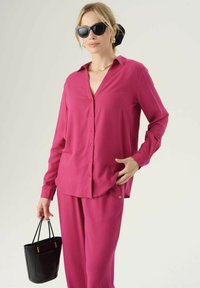 Blusa fucsia a maniche lunghe con chiusura a bottoni, abbinata a pantaloni coordinati. Una borsa nera strutturata e occhiali da sole completano il look.