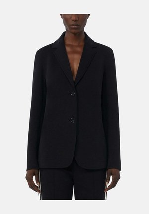 Vrouw draagt een zwarte tweeknopsblazer met inkepingrevers en bijpassende zwarte broek tegen een effen lichte achtergrond.