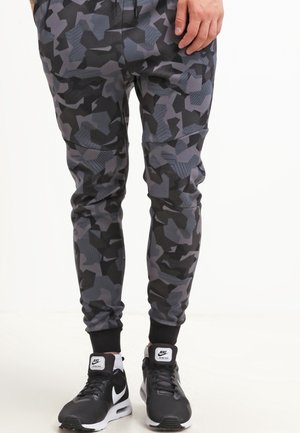 Graue und schwarze Camouflage-Jogginghosen mit gerippten Bündchen, Seitentaschen und flexibler Passform, kombiniert mit schwarz-weißen Nike-Sneakern.