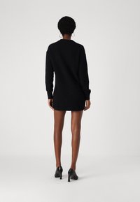 Robe noire en maille avec manches longues, col et poignets côtelés ; portée avec des chaussures à talons hautes noires. Texture lisse, longueur au-dessus du genou.