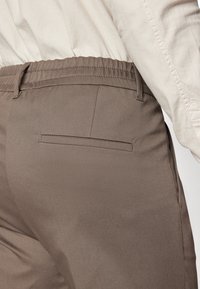 Pantalones de tela marrón con una textura suave, que presentan un bolsillo trasero y una cinturilla elástica. El material parece ligero y ajustado.