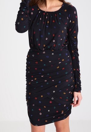Kobieta w dopasowanej czarnej sukience z długimi rękawami, ozdobionej drobnymi kolorowymi kółeczkami, marszczonej w talii i po bokach.