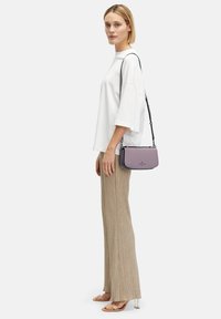 Fioletowa torebka o gładkiej fakturze, z zamkiem błyskawicznym i odpinanym paskiem, zestawiona z luźną białą koszulą oraz żeberkowanymi beżowymi spodniami.