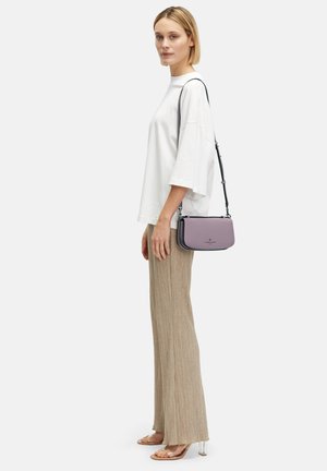 Fioletowa torebka o gładkiej fakturze, z zamkiem błyskawicznym i odpinanym paskiem, zestawiona z luźną białą koszulą oraz żeberkowanymi beżowymi spodniami.