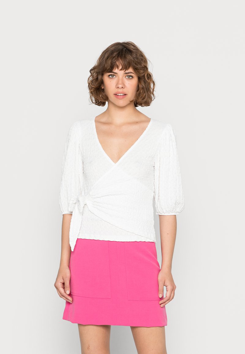 Blusa branca texturizada com decote em V e mangas bufantes, atada na cintura, combinada com uma saia rosa brilhante em linha A com bolsos.