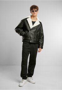 Jonge man in een zwart leren jack met een witte fleece kraag, zwarte broek, witte sneakers en een zonnebril, staand tegen een grijze achtergrond.