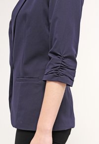 Granatowa marynarka z rękawami o długości siedmiu ósmych, z gromadzonymi detalami przy mankietach oraz jedną kieszenią z przodu, wykonana z gładkiego materiału.