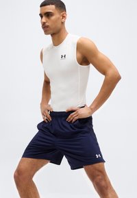 Άνδρας φορώντας λευκό αμάνικο συμπιεστικό μπλουζάκι και ναυτικό μπλε αθλητικό σορτς ποζάρει με τα χέρια στη μέση σε απλό φόντο.