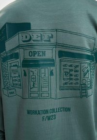 Graphic sweatshirt in muted green featuring a line-drawing storefront labeled "DEF" with "OPEN" sign and details of products displayed.