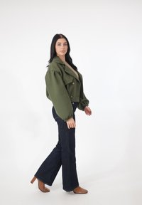 Oliwkowo-zielona krótka kurtka z nadmiernie szerokimi rękawami, złote guziki, w połączeniu z ciemnymi dzwonowatymi jeansami i brązowymi, szpiczastymi obcasami, na białym tle.