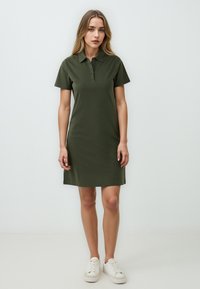 Sukienka polo w oliwkowym kolorze, z krótkimi rękawami, z zapięciem na guziki, długości do kolan, wykonana z gładkiego materiału, zestawiona z białymi sneakersami.