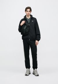 Μαύρο μπουφάν με φερμουάρ και ψηλό γιακά, σε συνδυασμό με μαύρο παντελόνι φόρμας με μπεζ ρίγες στο πλάι. Γκρι αθλητικά παπούτσια και μαύρη τσάντα ώμου.