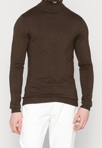 Maglione a collo alto marrone realizzato in tessuto morbido, con maniche lunghe e polsini e orlo a coste, abbinato a pantaloni bianchi.