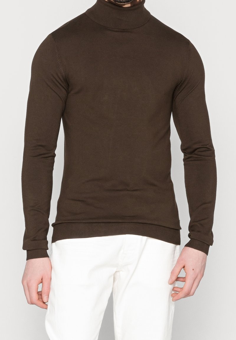 Maglione a collo alto marrone realizzato in tessuto morbido, con maniche lunghe e polsini e orlo a coste, abbinato a pantaloni bianchi.