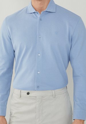 Chemise bleu clair à manches longues, faite en tissu lisse. Présente un logo discret, un col arrondi et six boutons assortis.