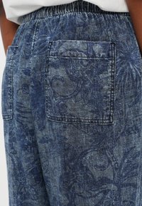 Blue patterned denim trousers feature an elastic waistband, two side pockets, and a textured floral design in varying blue tones.