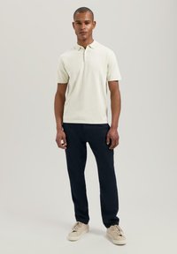 Kortermet polo i off-white bomull med krage og to-knapps placket, paret med mørk marineblå bukser og beige joggesko.