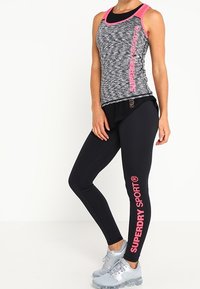 Zwart en grijs gemarmerde tanktop met roze accenten, gecombineerd met zwarte leggings met een roze "SUPERDRY SPORT" logo, en lichtgrijze sportschoenen.