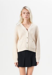 Beige gebreide cardigan met een V-hals, knoopsluiting aan de voorkant en twee opgestikte zakken; heeft goudkleurige knopen en ribgebreide manchet.