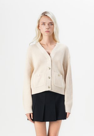 Beige gestrickte Strickjacke mit V-Ausschnitt, Druckknopfverschluss und zwei aufgenähten Taschen; mit goldfarbenen Knöpfen und gerippten Bündchen.