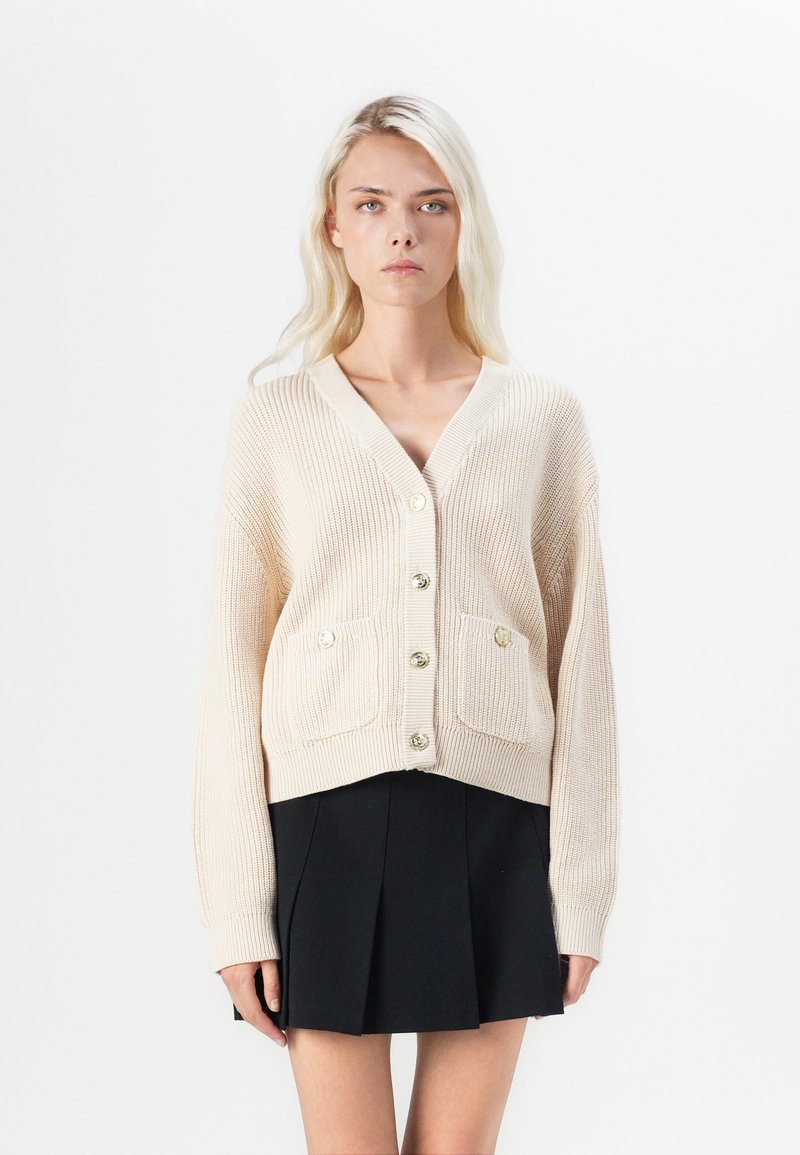 Beige gebreide cardigan met een V-hals, knoopsluiting aan de voorkant en twee opgestikte zakken; heeft goudkleurige knopen en ribgebreide manchet.